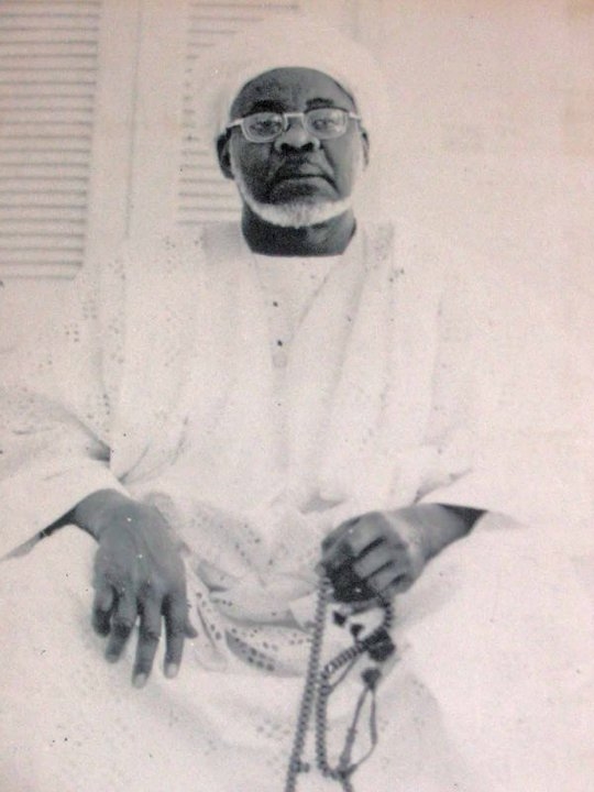 Portrait of Shaykh Ibrāhīm Niasse (Baye Niasse) seated in white robes holding prayer beads